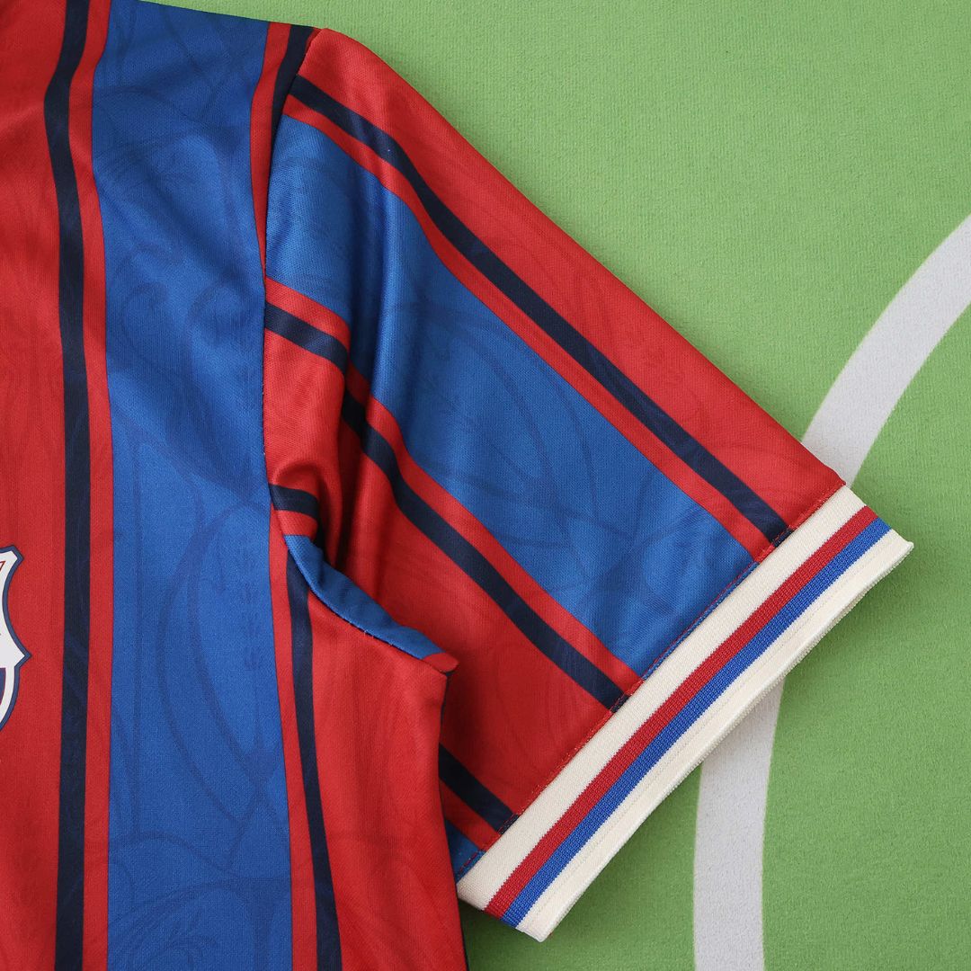 Camisola de futebol Barcelona com detalhes em azul e vermelho, disponível na OLYMPIC STORE.