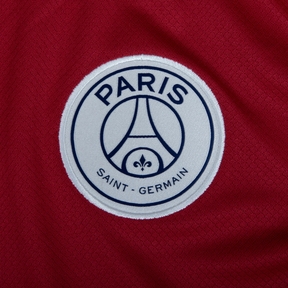 Emblema do PSG em camisola de futebol vermelha, representando o Paris Saint-Germain.