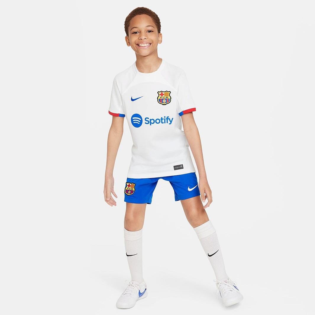 Conjunto Infantil Nike FC Barcelona 2023/24 II, menino usando camisa e short do time em cores brancas e azuis.