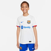 Conjunto Infantil Nike FC Barcelona 2023/24 II, menino sorridente usando uniforme do time, camiseta branca e short azul.