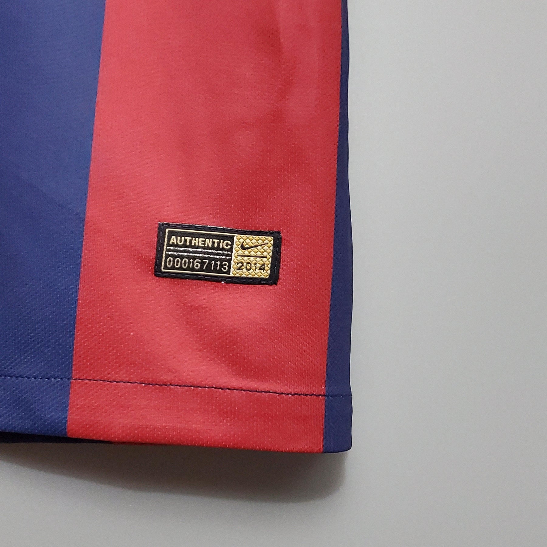 Detalhe do selo autenticidade da camiseta Barcelona Home 14/15 Retro Olympic Store com cores vermelha e azul.