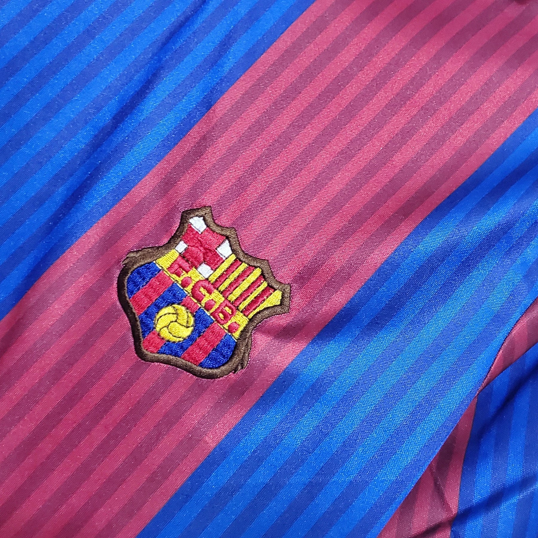 Emblema do FC Barcelona em camisa listrada das cores azul e grená, representando o Barcelona.