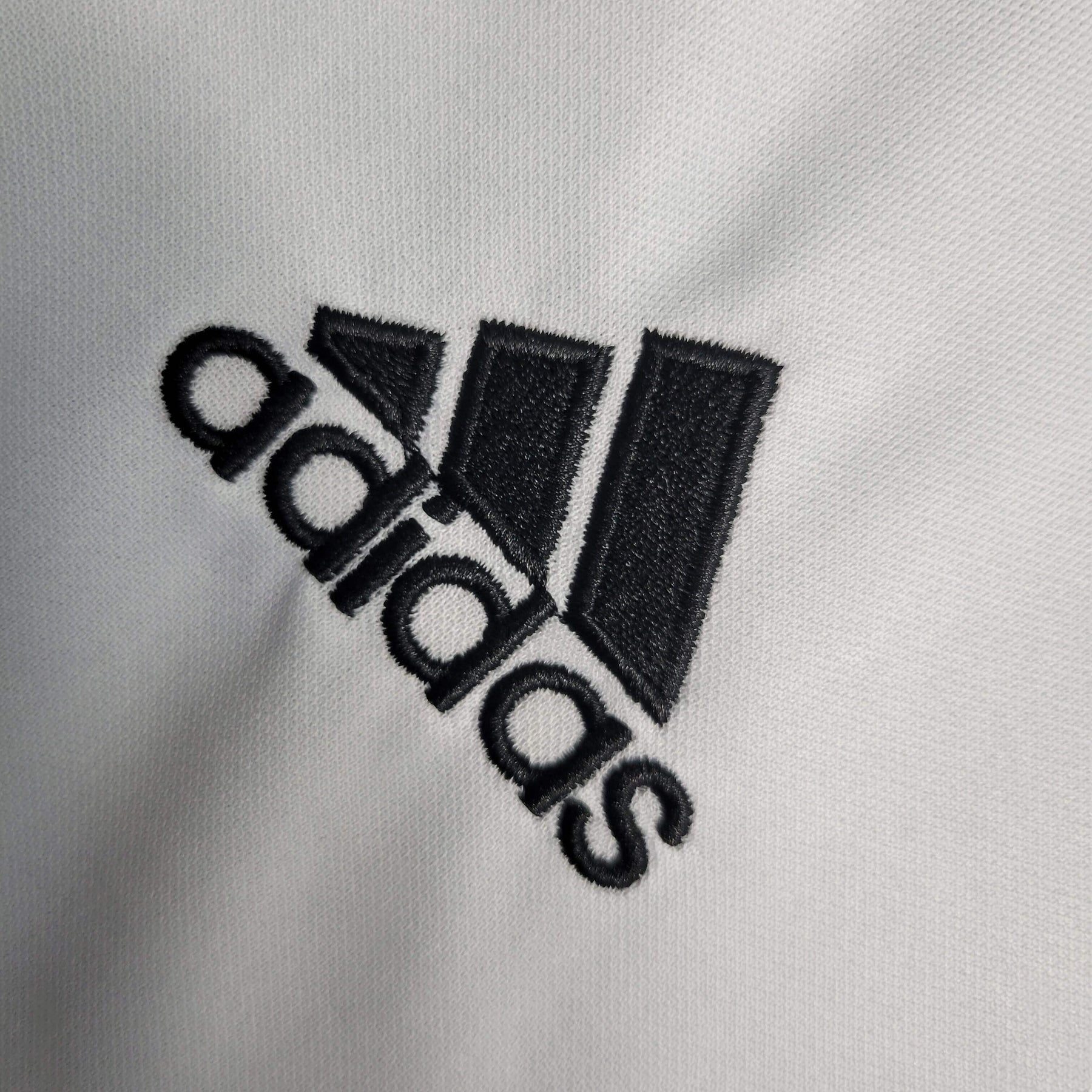 Logo da marca adidas em destaque, bordado em preto sobre tecido claro.