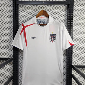 Camisola Inglaterra home 2005 Retro exposta em um cabide sobre uma grade de metal.